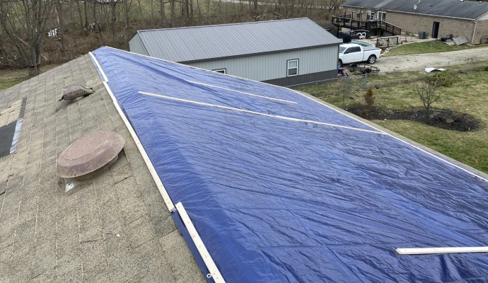 A large blue emergency tarp secured with wooden strips over a damaged residential shingle roof