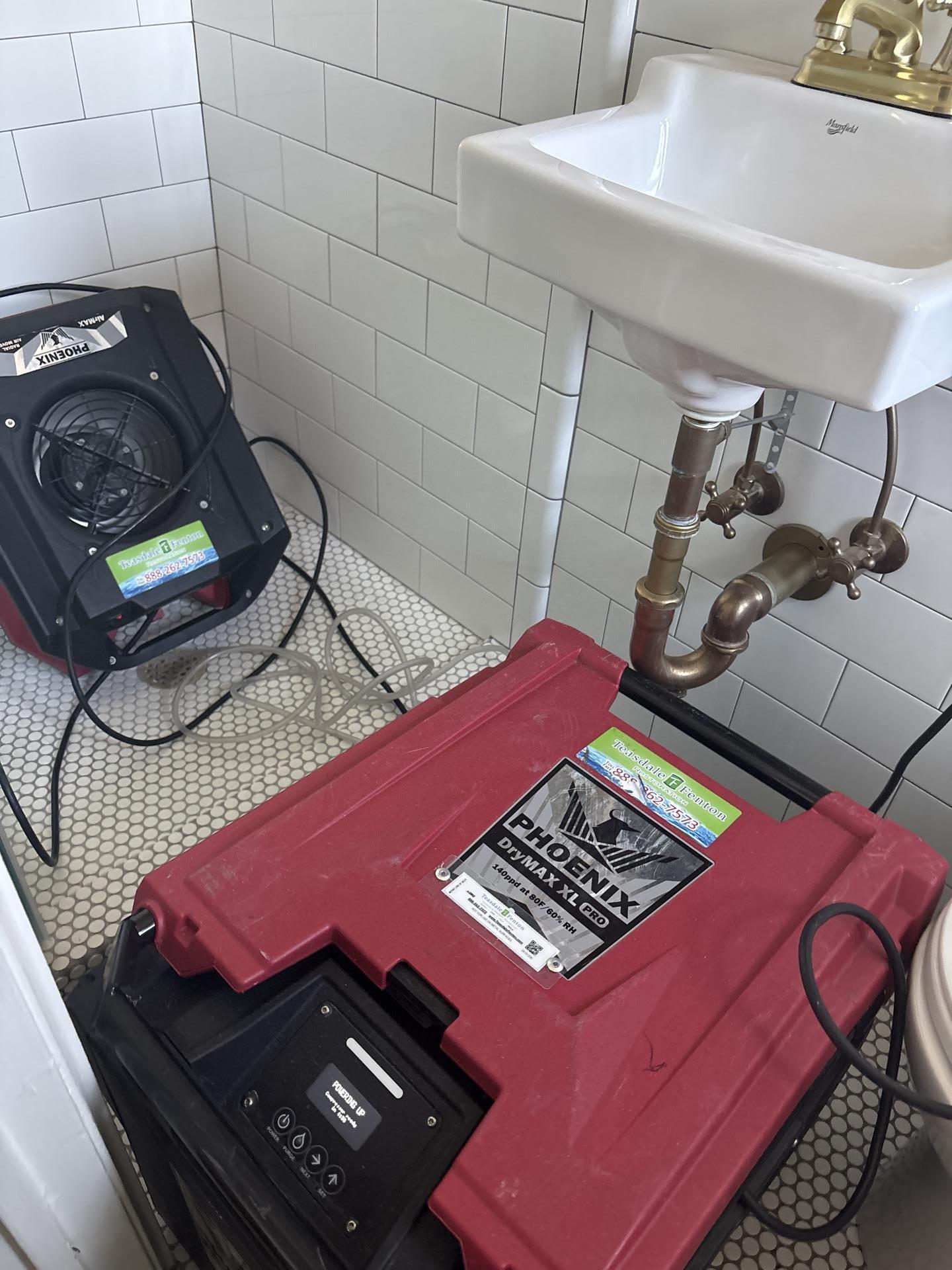 Professional Phoenix drying equipment, including a red dehumidifier and a black air mover, set up in a white-tiled bathroom for effective water damage restoration by Teasdale Fenton.