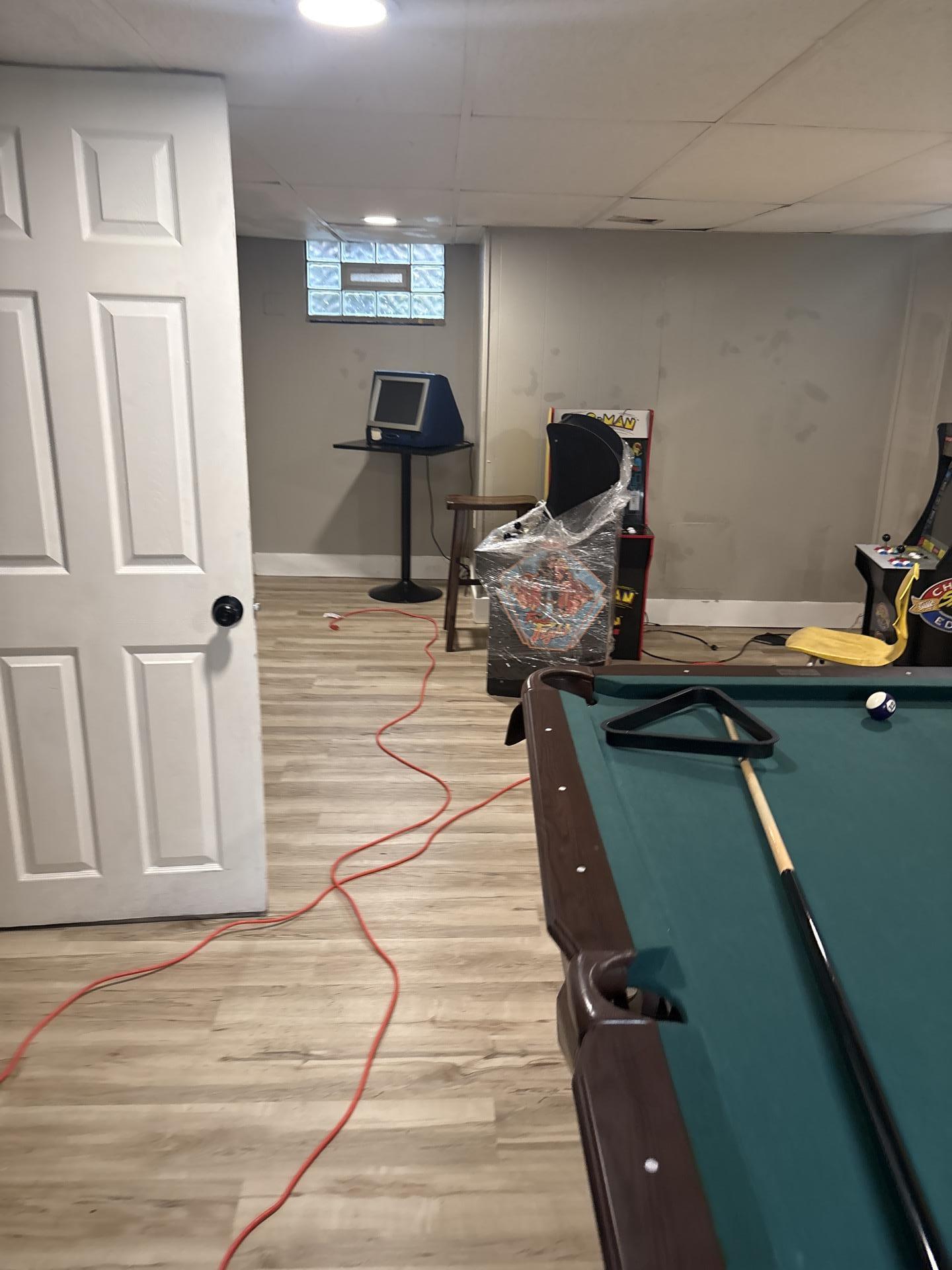Basement recreation room showing water damage and active remediation equipment including a long red power cord and drying fans on laminate flooring.