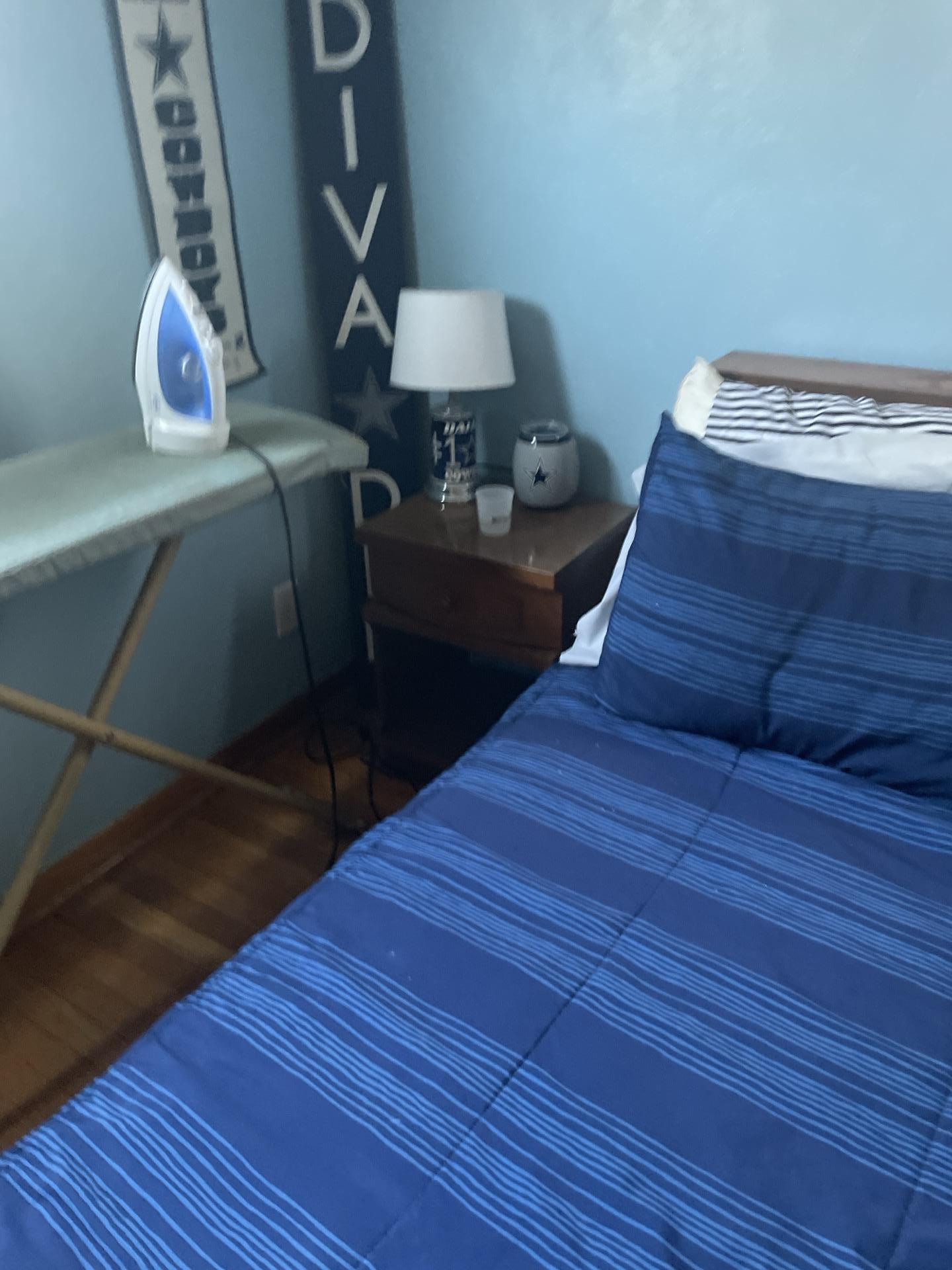 A bedroom interior featuring an ironing board with an iron, a nightstand with a lamp, and a bed with blue striped bedding.
