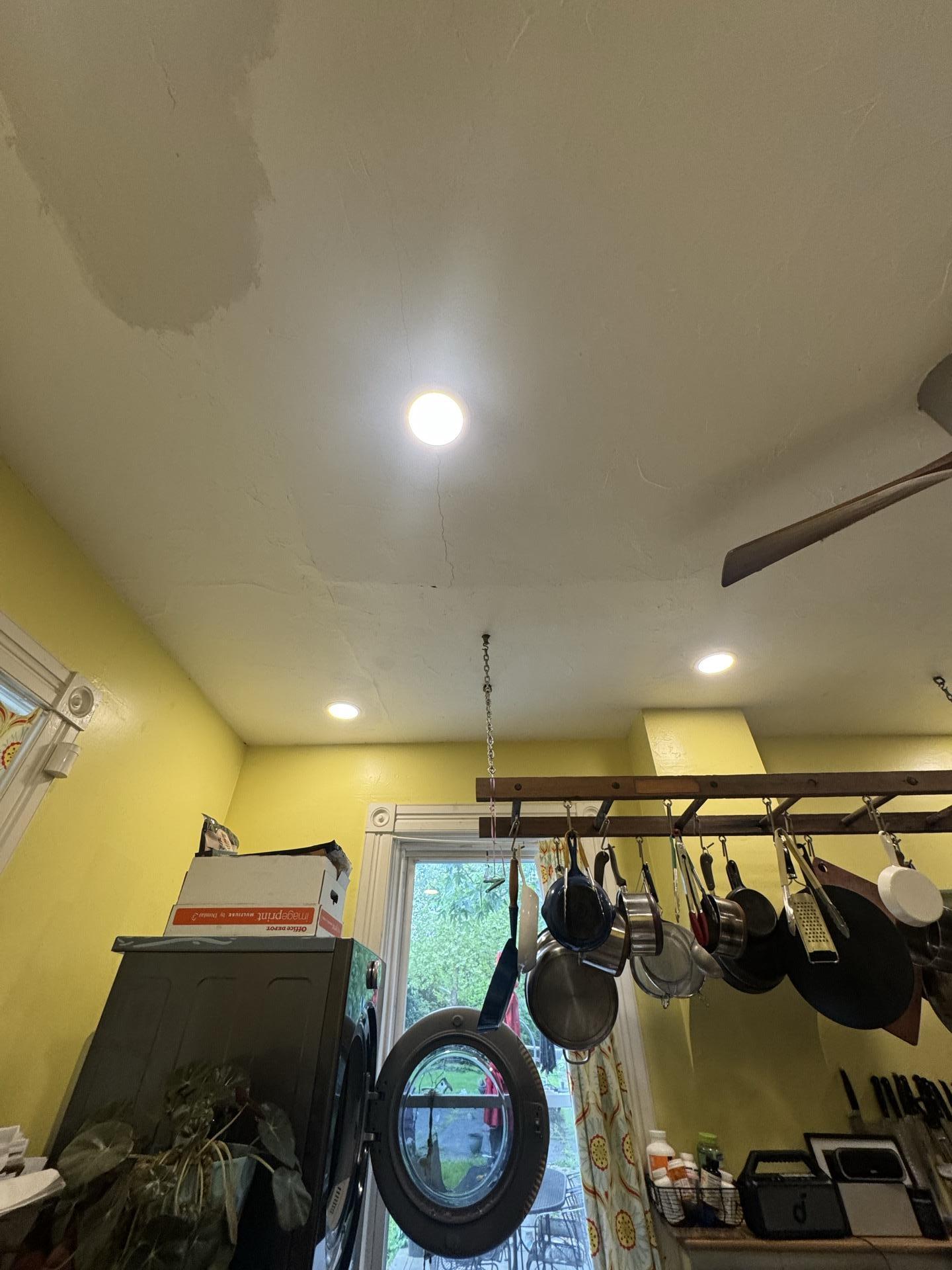 Wider shot of a kitchen ceiling with visible water stains, cracks, and peeling paint, indicating significant water damage above a washing machine and pot rack.