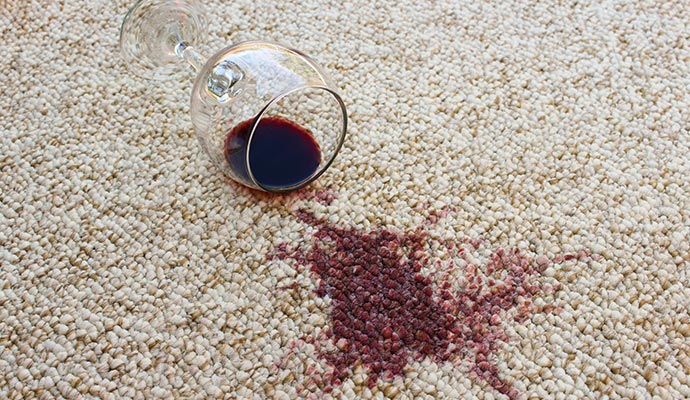 A clear glass of red wine is lying on its side on a light-colored carpet with a dark red wine stain spilling out