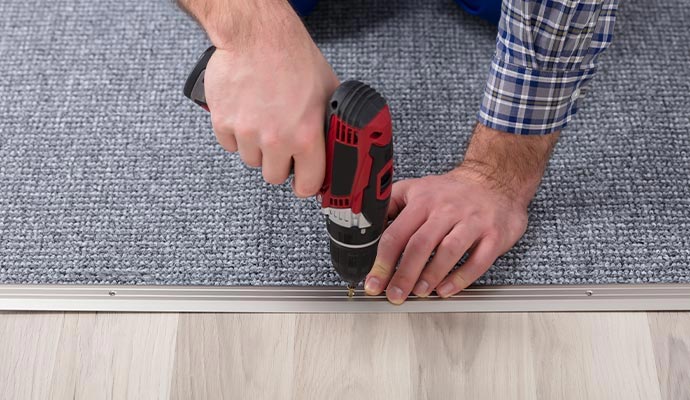 A technician using a power drill to install a metal transition strip between grey carpet and light hardwood flooring